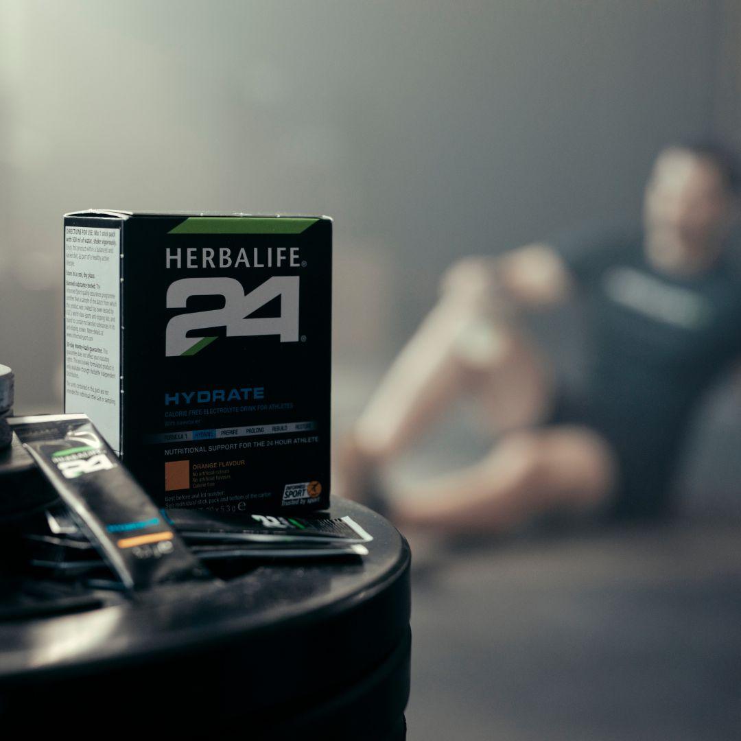 En pakke Herbalife H24 Hydrate isotonisk sportsdrikk og to løse porsjonsposer står på en stabel vektskiver. Utydelig i bakgrunnen ses en mann i treningstøy.