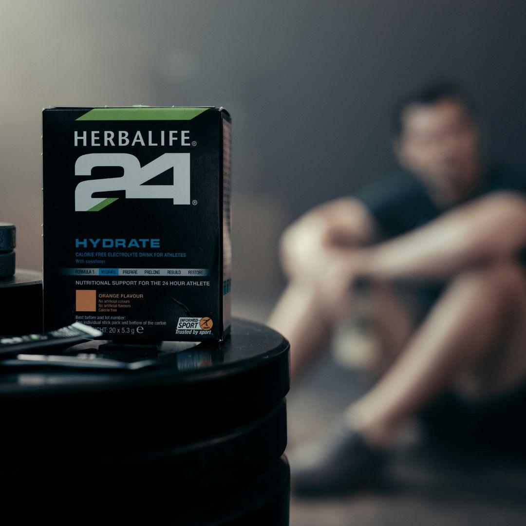 En pakke Herbalife H24 Hydrate isotonisk sportsdrikk og noen løse porsjonsposer står på en stabel vektskiver. Utydelig i bakgrunnen skimtes en mann i treningstøy.