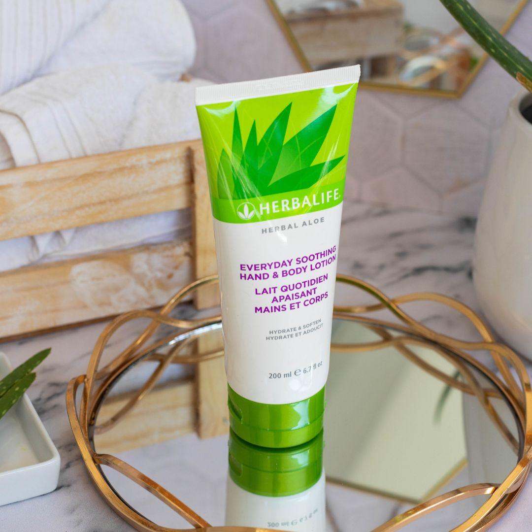 Tube med Herbalife Herbal Aloe Everyday Soothing Hand & Body Lotion på glassfat med gullkant på marmorbord. Aloe vera-plante, oppskårne aloevera-blad og trekasse med håndklær i bakgrunnen.