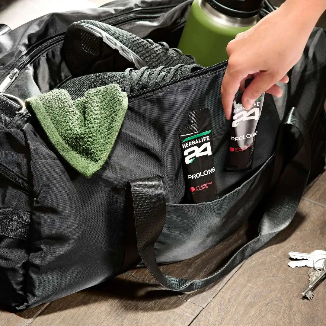 En åpen treningsbag med sko og drikkeflaske og en hånd som tar en av to poser Herbalife H24 Prolong Energy Gel fra den.