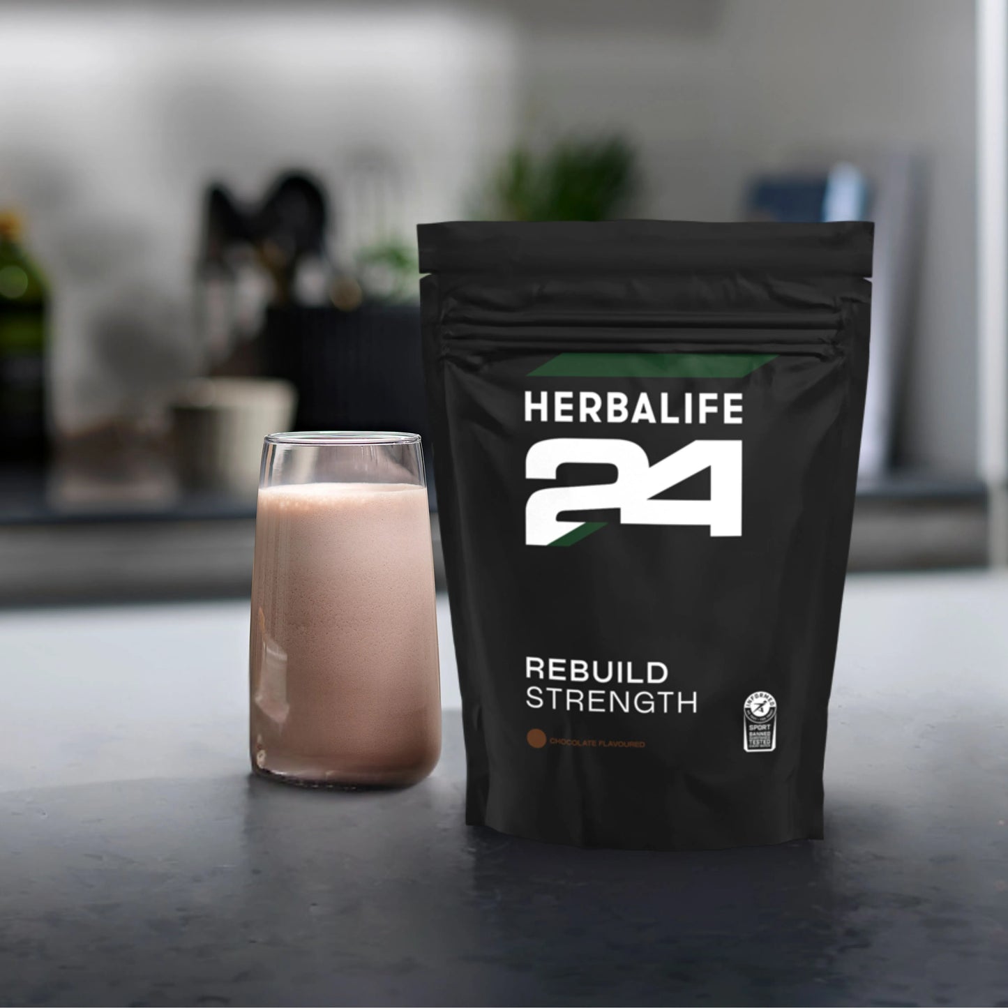 H24 Rebuild Strength pose med et glass proteinshake på en kjøkkenbenk.