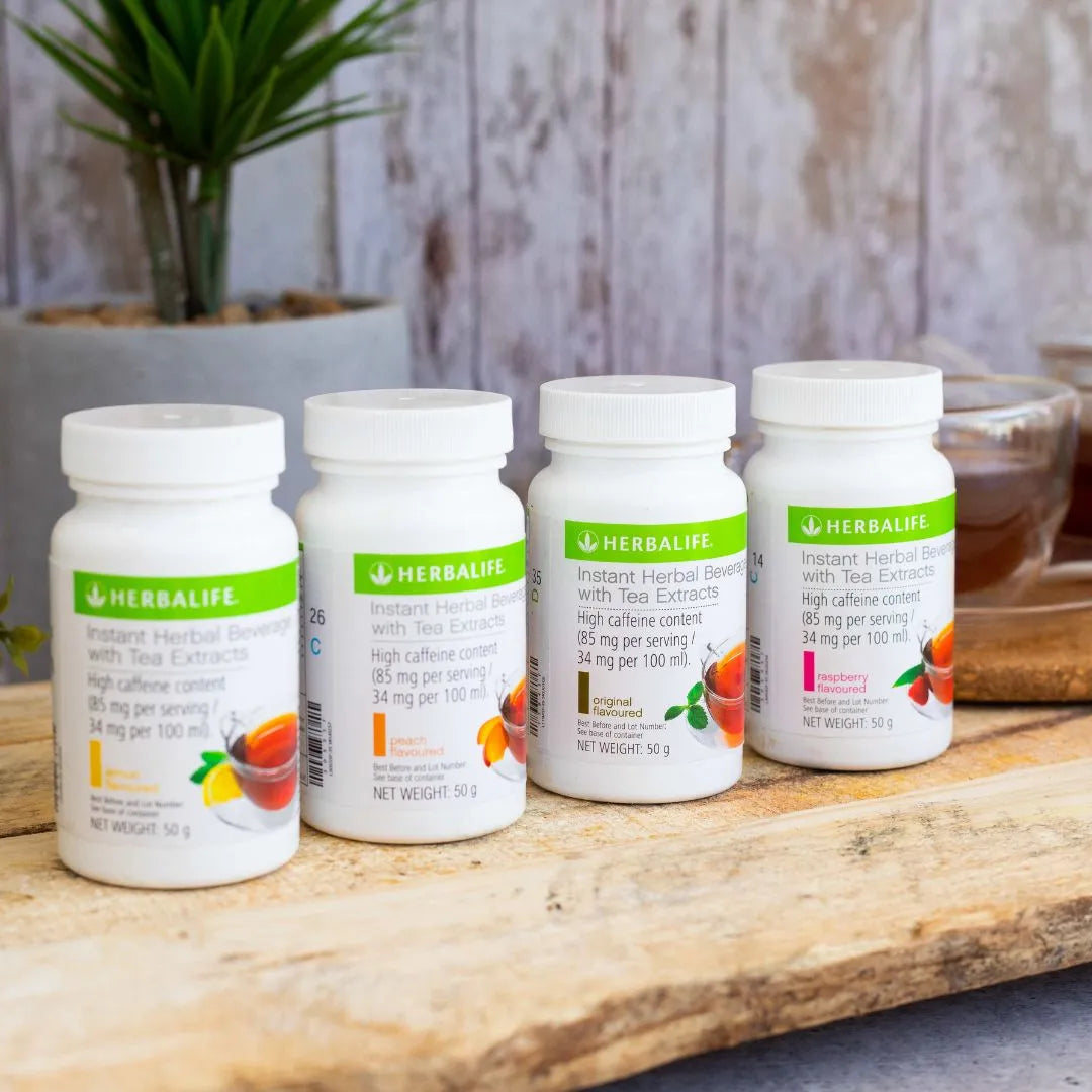 Fire bokser Herbalife Instant urtekonsentrat te med de ulike smakene sitron, fersken, original og bringebær på en treplanke. I bakgrunnen skimtes en kopp urtete.