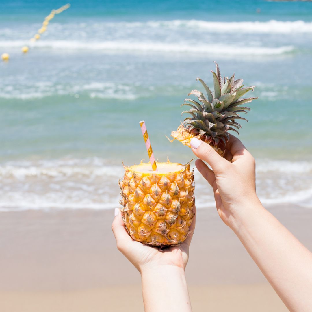 Herbalife Formula 1 shake Pineapple Coconut Limited Edition servert i en uthult ananas med sugerør, holdt i hendene foran stranden og havet.