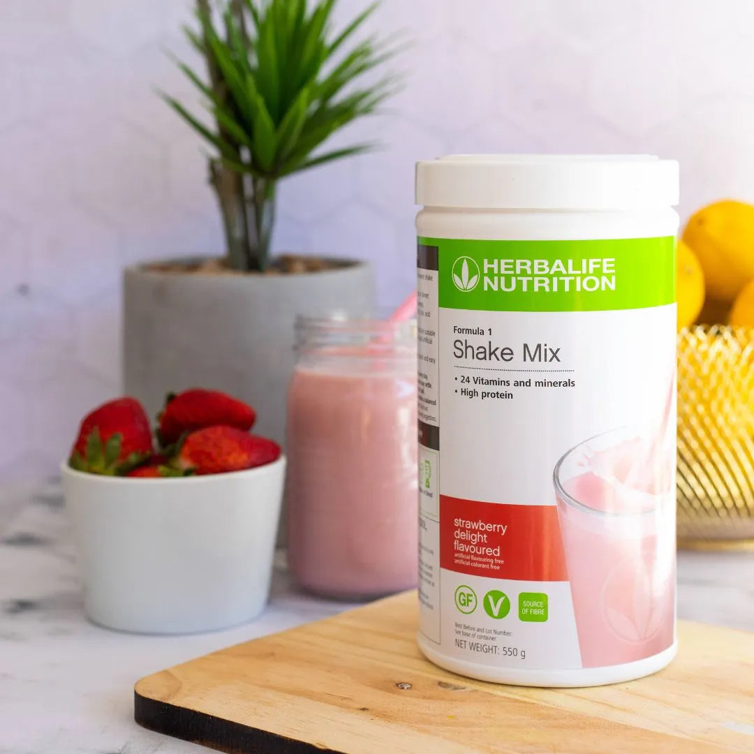 En boks Herbalife jordbær-shake - Formula 1 shakemix med Strawberry Delight-smak - på trefjøl på marmorbord. I bakgrunnen er en glasskål med gule sitroner, en jordbærshake i et glasskrus med rosa sugerør, en skål med røde jordbær og en grønn plante.