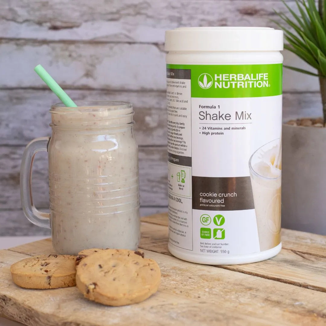 Herbalife Formula 1 shakemix med Cookie Crunch-smak og en smoothie på ei lys trefjøl. Smoothien serveres med sugerør i et glasskrus. I forgrunnen tre cookies med sjokoladebiter.