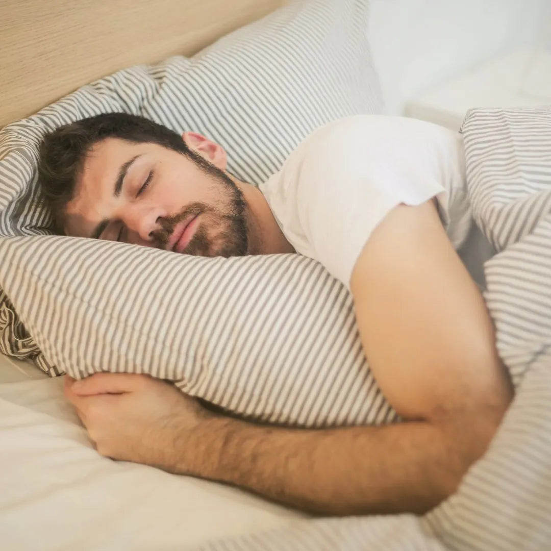 En mann med skjegg og hvit t-skjorte sover i stripete sengetøy.