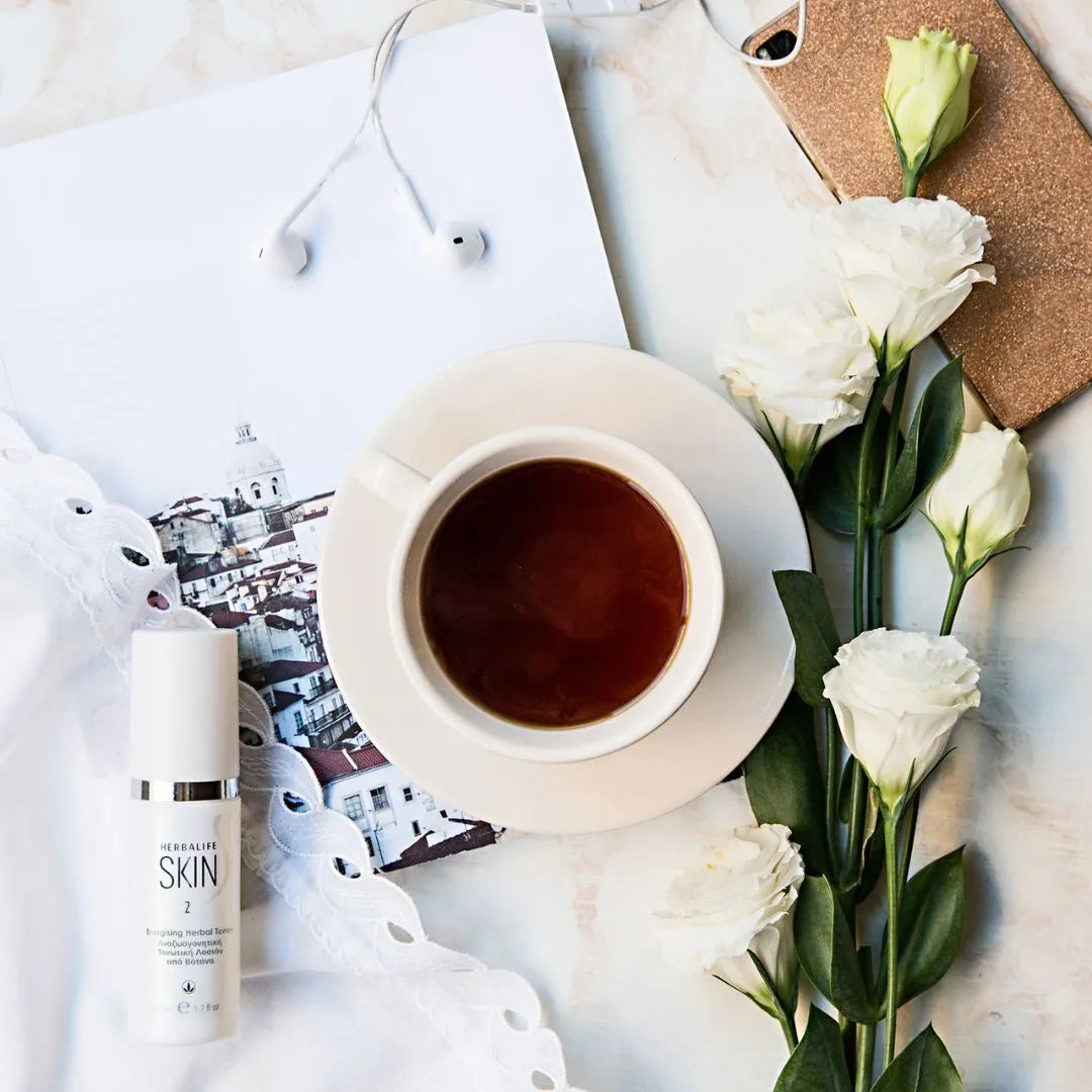 En sprayflaske Herbalife SKIN Energising Herbal Toner ansiktsvann på et bord sammen med en kopp te, en mobiltelefon med headset og noen hvite blomster.