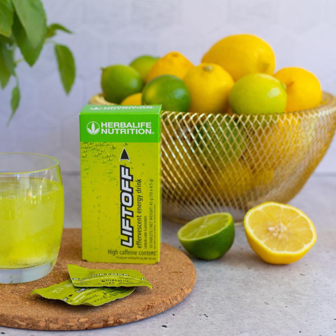 En pakke Herbalife Liftoff energidrikk står sammen med et glass liftoff, to løse porsjonspakker og en skål med lime og sitroner.