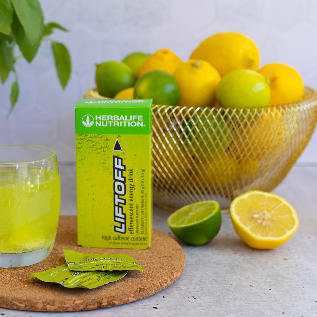 En pakke Herbalife Liftoff energidrikk står sammen med et glass Liftoff, to løse porsjonspakker og en skål med lime og sitroner.