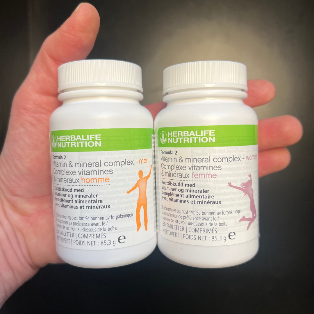 Multivitamin Herbalife Formula 2, en versjon for kvinne og en for mann i hånda. Boksene har påskriften Kosttilskudd med vitaminer og mineraler.