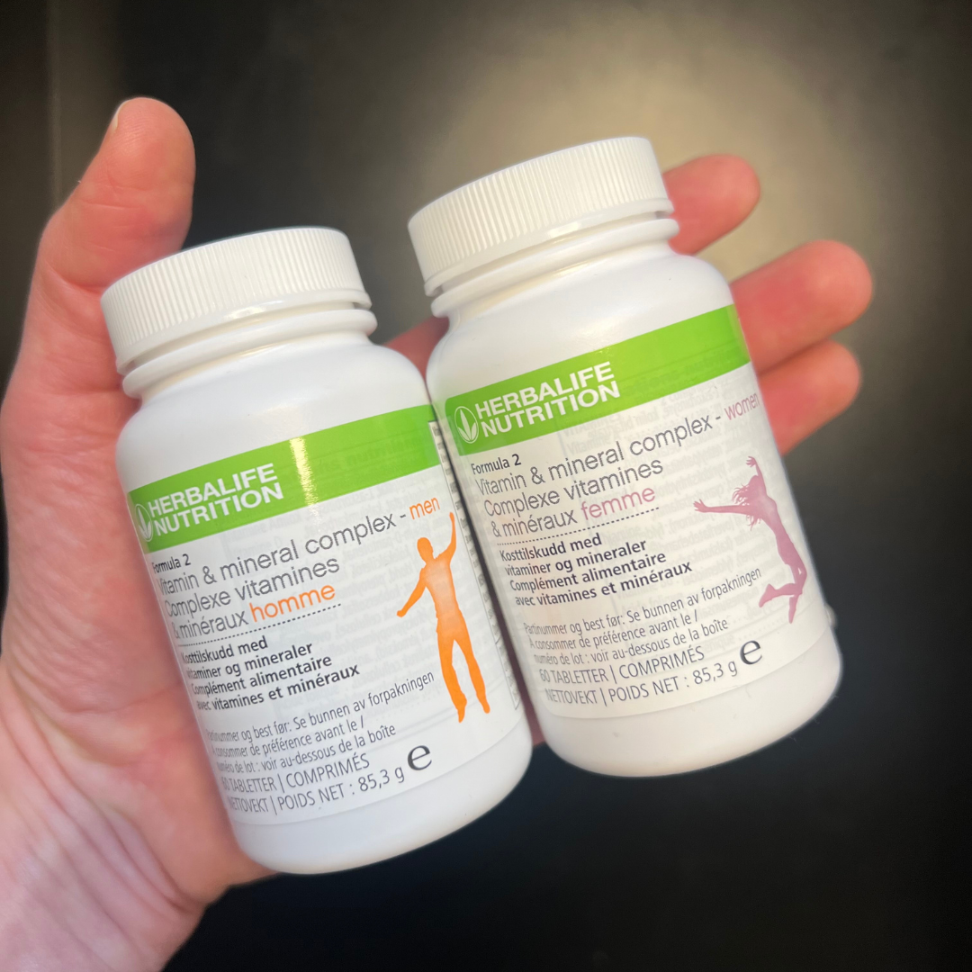 Multivitamin Herbalife Formula 2, en versjon for kvinne og en for mann på skrå i hånda. Boksene har påskriften Kosttilskudd med vitaminer og mineraler.