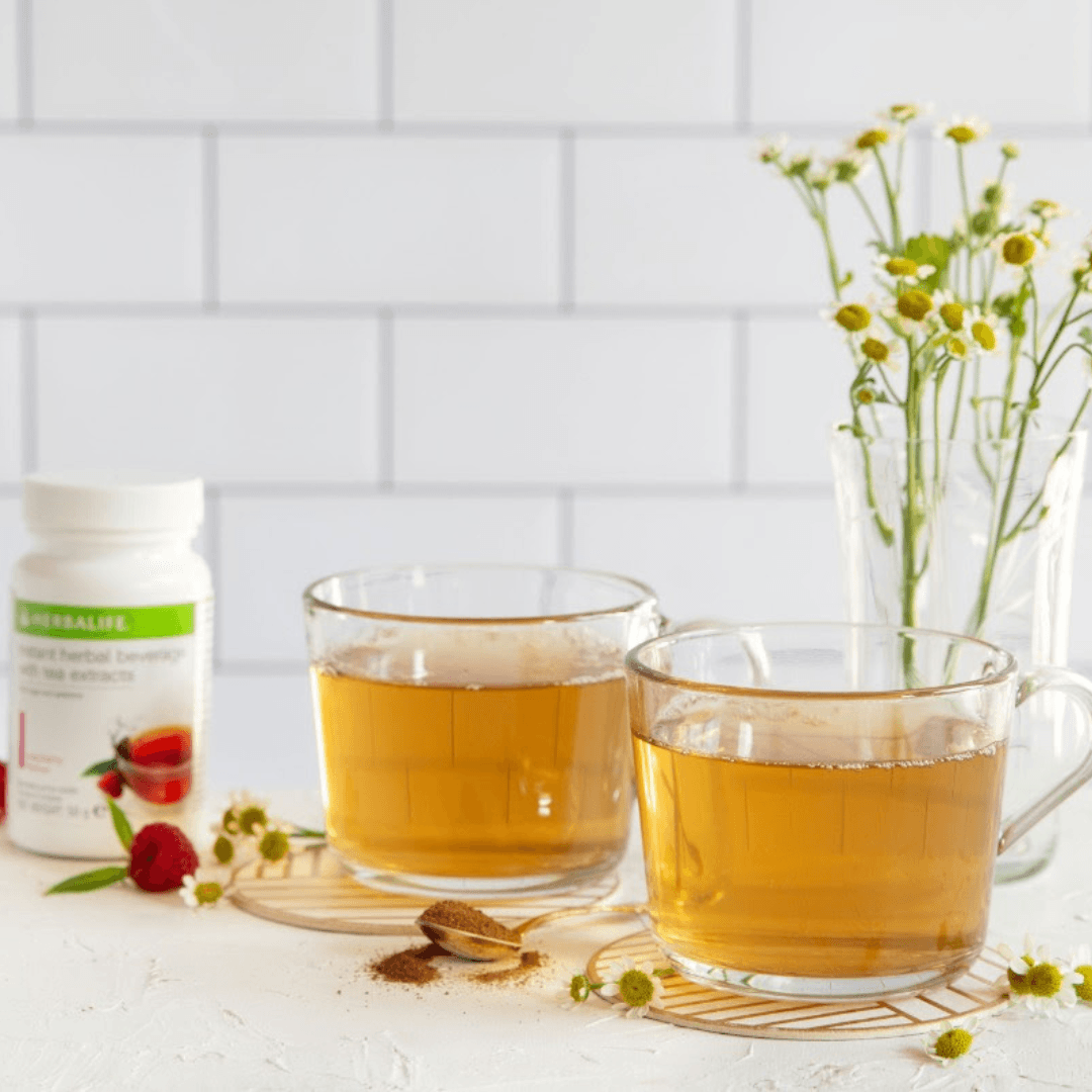 To glasskopper bringebær-te foran en vase med hestehov, et ferskt bringebær på bordet ligger sammen med en teskje instant te. I bakgrunnen ses en boks Herbalife urtekonsentrat raspberry.