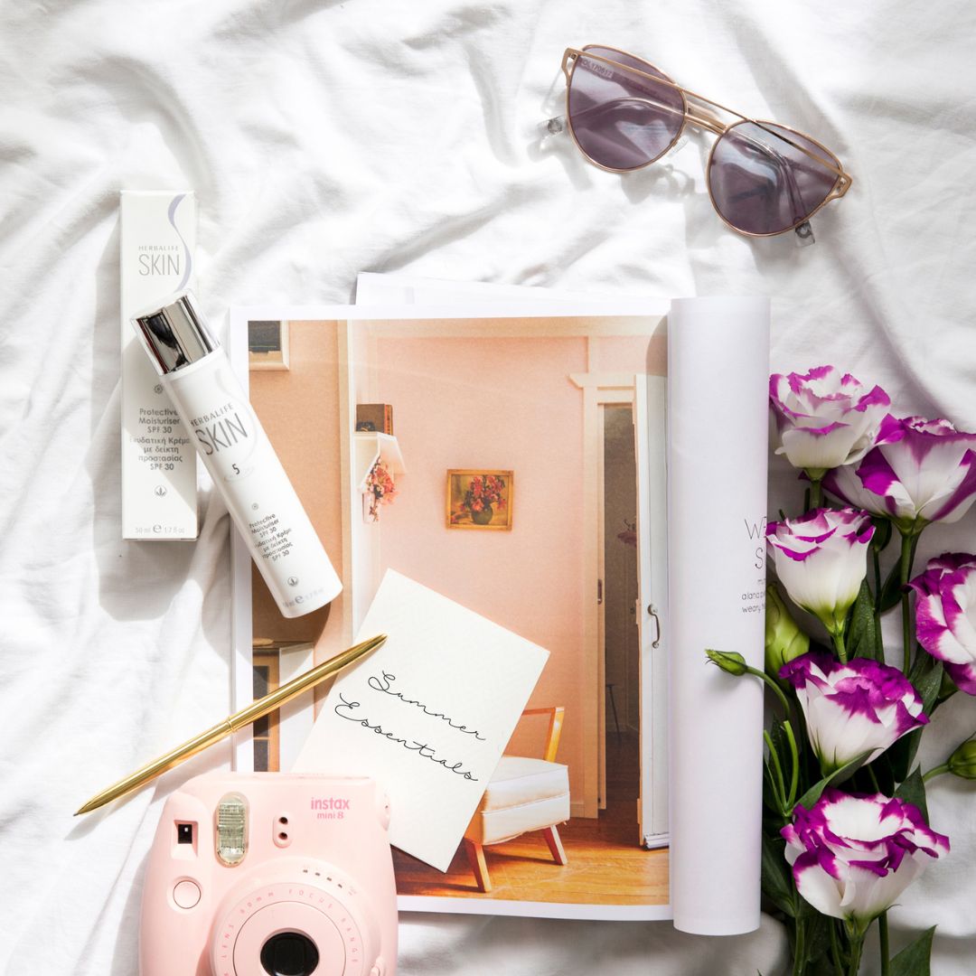 En tube Herbalife SKIN Protective Moisturiser dagkrem med solfaktor 30 ligger sammen med sommerrelaterte ting, som solbriller, kamera, blomster og et magasin samt en lapp med teksten “Summer Essentials”.