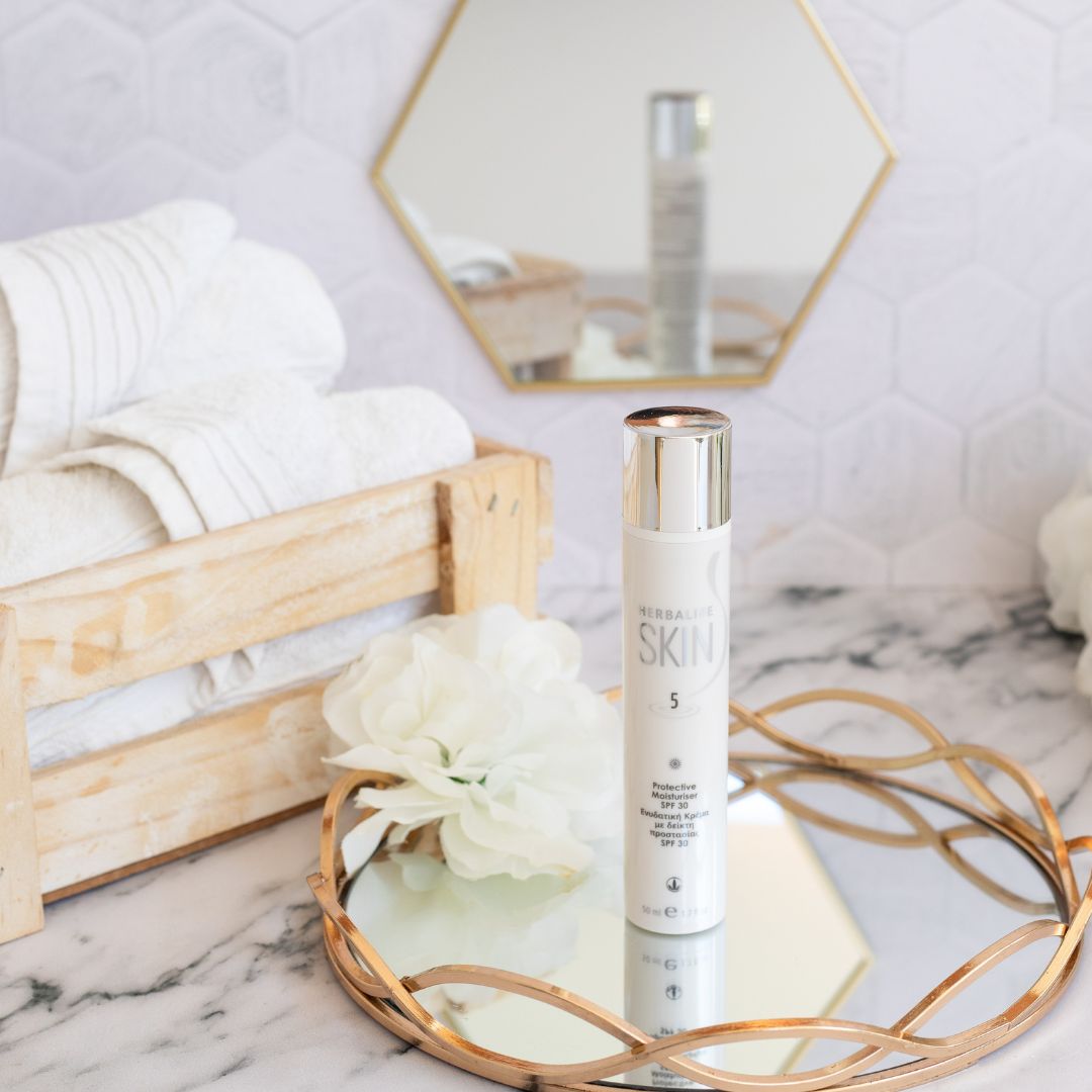 En tube Herbalife SKIN Protective Moisturiser dagkrem med solfaktor 30 står på et glassfat med gullkant. Hvite håndklær ligger i en trekasse i bakgrunnen.