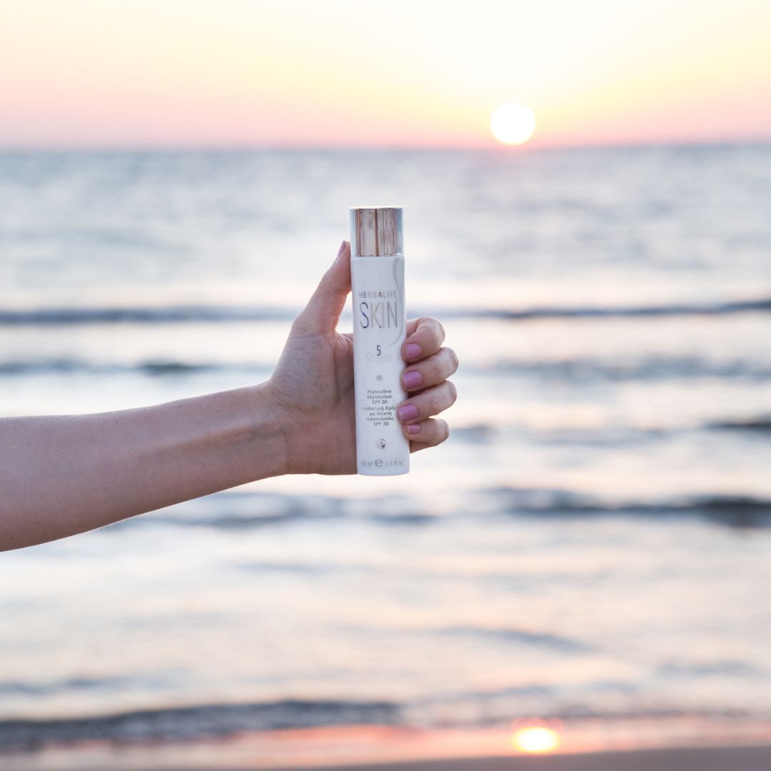 En hånd holder en tube Herbalife SKIN Protective Moisturiser dagkrem med solfaktor 30, med havet i solnedgang i bakgrunnen.