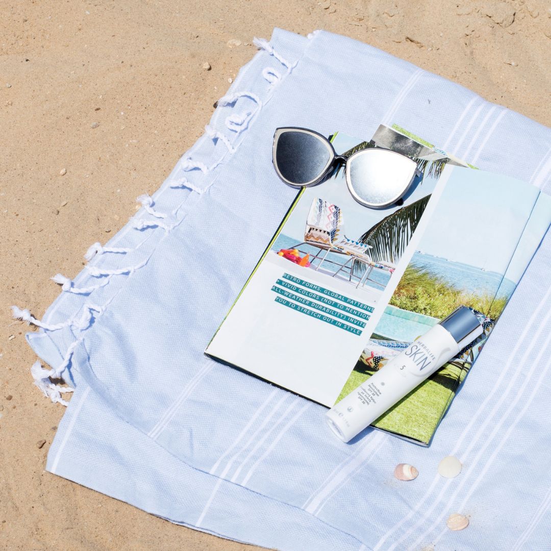 En tube Herbalife SKIN Protective Moisturiser dagkrem med solfaktor 30 ligger sammen med et reisemagasin og et par solbriller på et håndkle på en strand.