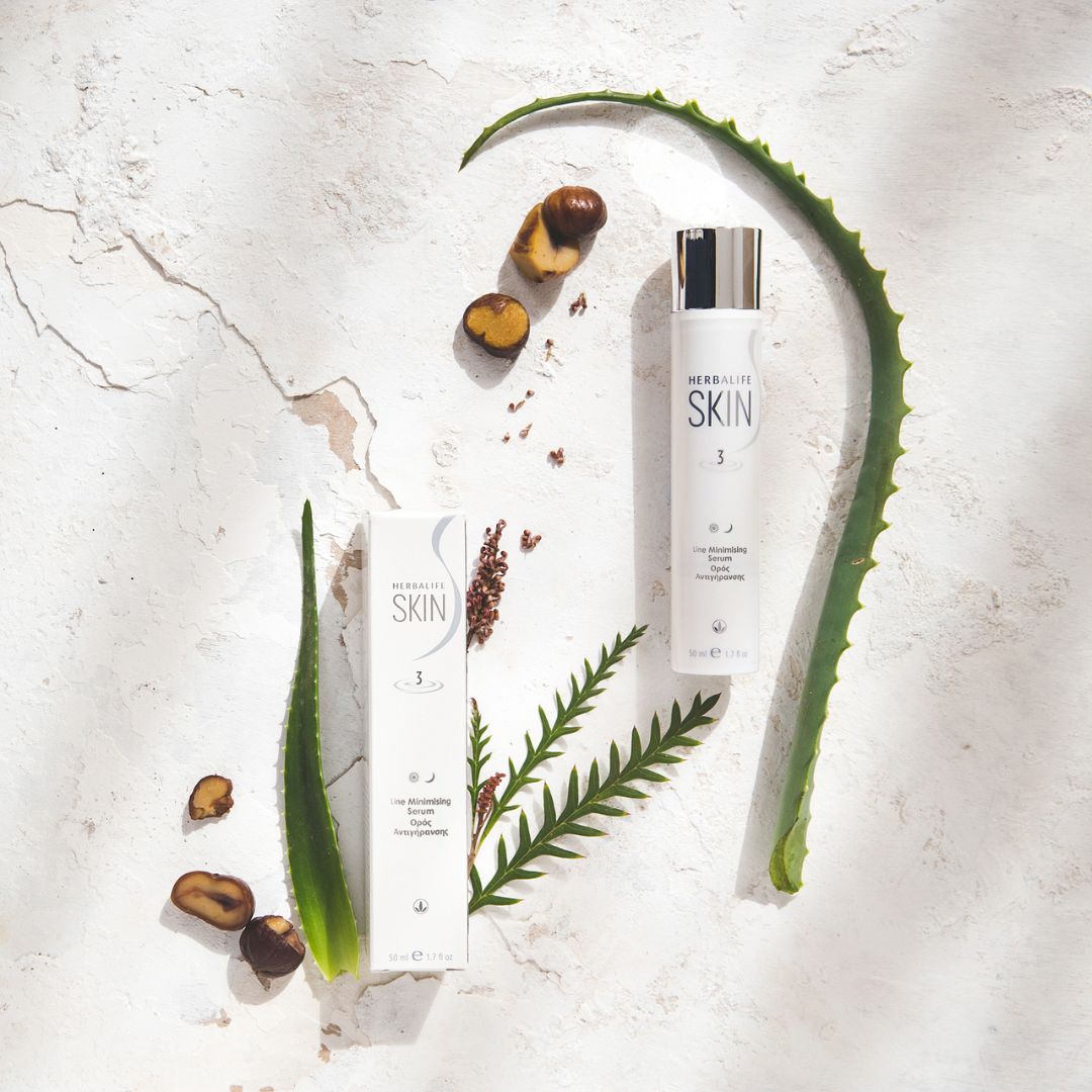 En tube og en eske Herbalife SKIN Line Minimising Serum ligger på et bord sammen med aloeblader og noen nøtter.