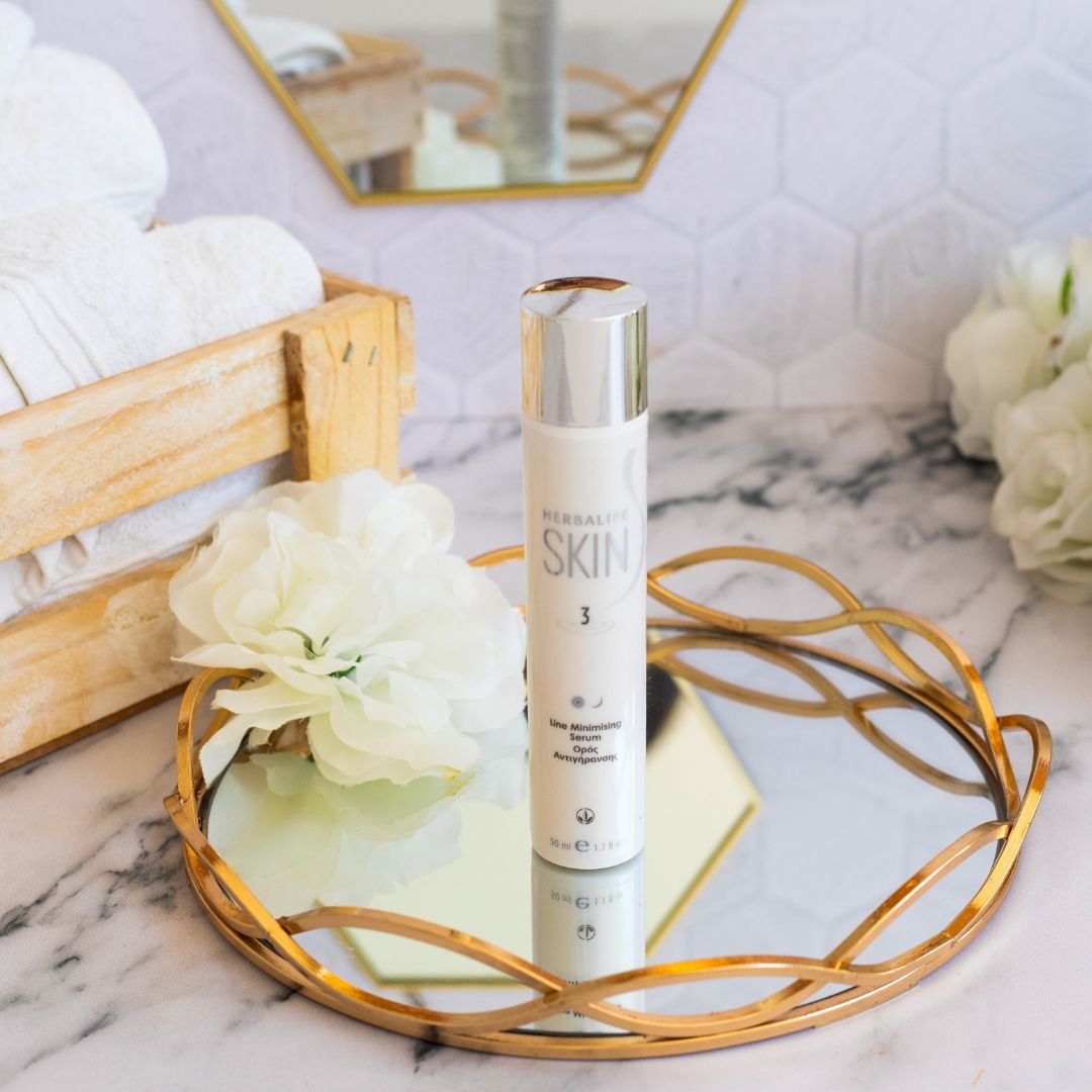 En tube Herbalife SKIN Line Minimising Serum står på et glassfat med gullkant. Hvite håndklær ligger i en trekasse i bakgrunnen.