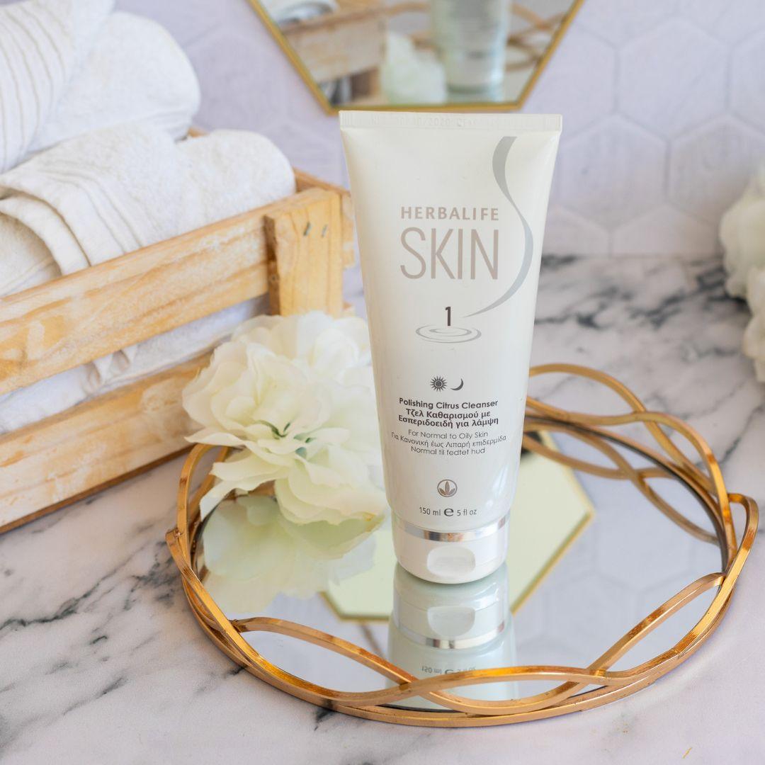En tube Herbalife SKIN Citrus Cleanser på et glassfat med gullkant. Noen sammenrullede håndklær i en trekasse bak.