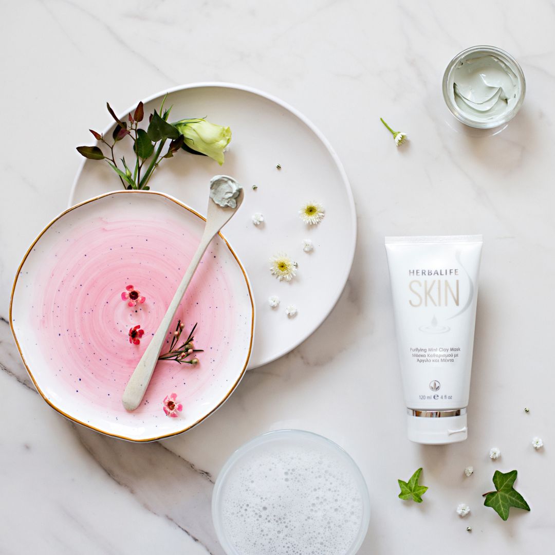 En tube Herbalife SKIN Purifying Mint Clay Mask ansiktsmaske ligger på et bord ved siden av et par asjetter med blomster og en skje med grønn ansiktsmaske.