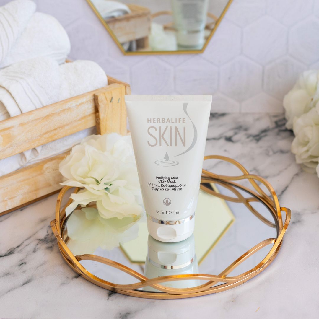 En tube Herbalife SKIN Purifying Mint Clay Mask grønn ansiktsmaske står på et glassfat med gullkant. I bakgrunnen ligger hvite håndklær i en trekasse.