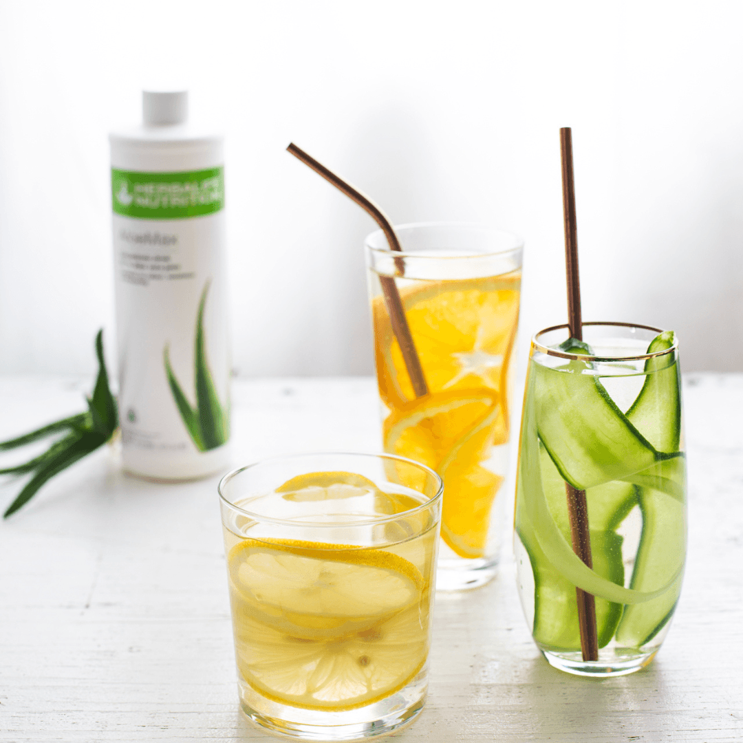 Tre glass vann med aloevera-juice og skivet agurk, appelsin og sitron. I bakgrunnen skimtes ei flaske Herbalife Herbal Aloe AloeMax urtekonsentrat.