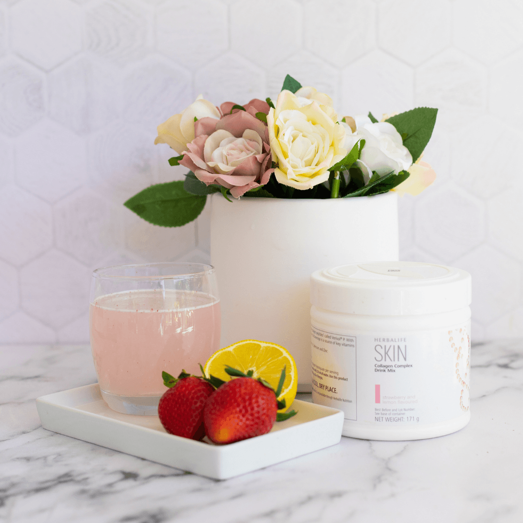 En boks Herbalife SKIN Collagen Booster står på et bord sammen med et glass drikkeklar kollagen, to jordbær og en halv sitron. Bak står en blomstervase.