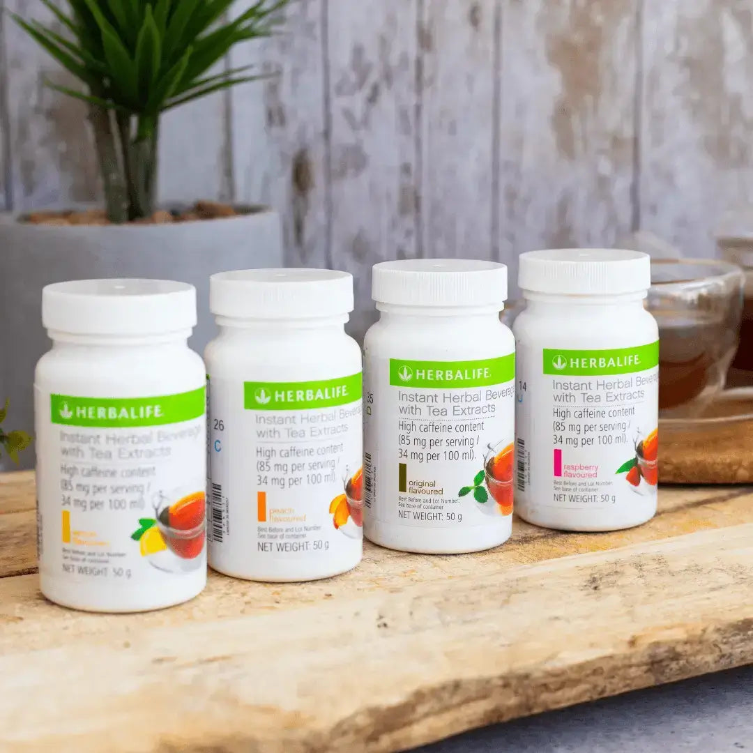 Fire bokser Herbalife Instant urtekonsentrat te med de ulike smakene sitron, fersken, original og bringebær på en treplanke. I bakgrunnen skimtes en kopp urtete.