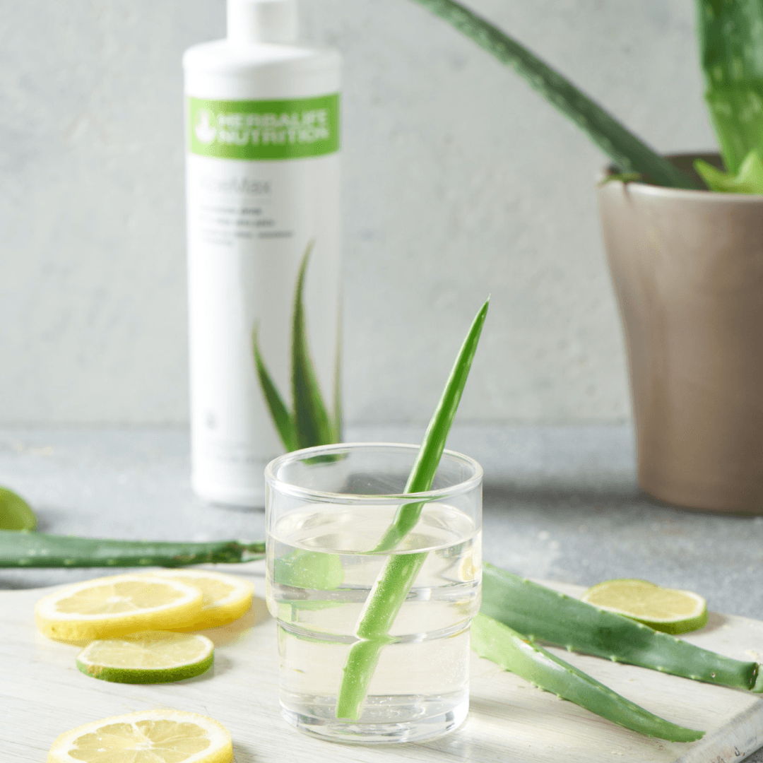 I forgrunnen et glass vann med aloevera-blad i. Aloe vera-blader og skivet sitron og lime ligger på bordet. I bakgrunnen skimtes en flaske Herbalife Herbal Aloe AloeMax urtekonsentrat aloejuice.