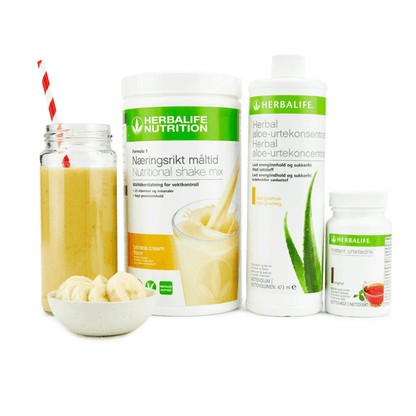 Bildet viser Herbalife-produktene som Frokostpakke basic banan består av; Herbalife-shake med banansmak, Herbal Aloe aloevera-juice med mangosmak og Instant urtete med original-smak samt en banan-shake i et glass med sugerør og noen bananskiver i en skål.