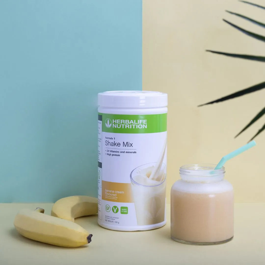 Boks Herbalife Formula 1 shakemix med banana cream-smak, et glasskrus med banan-shake og mintgrønt sugerør. To uskrelte gule bananer og en grønn plante i bakgrunnen.