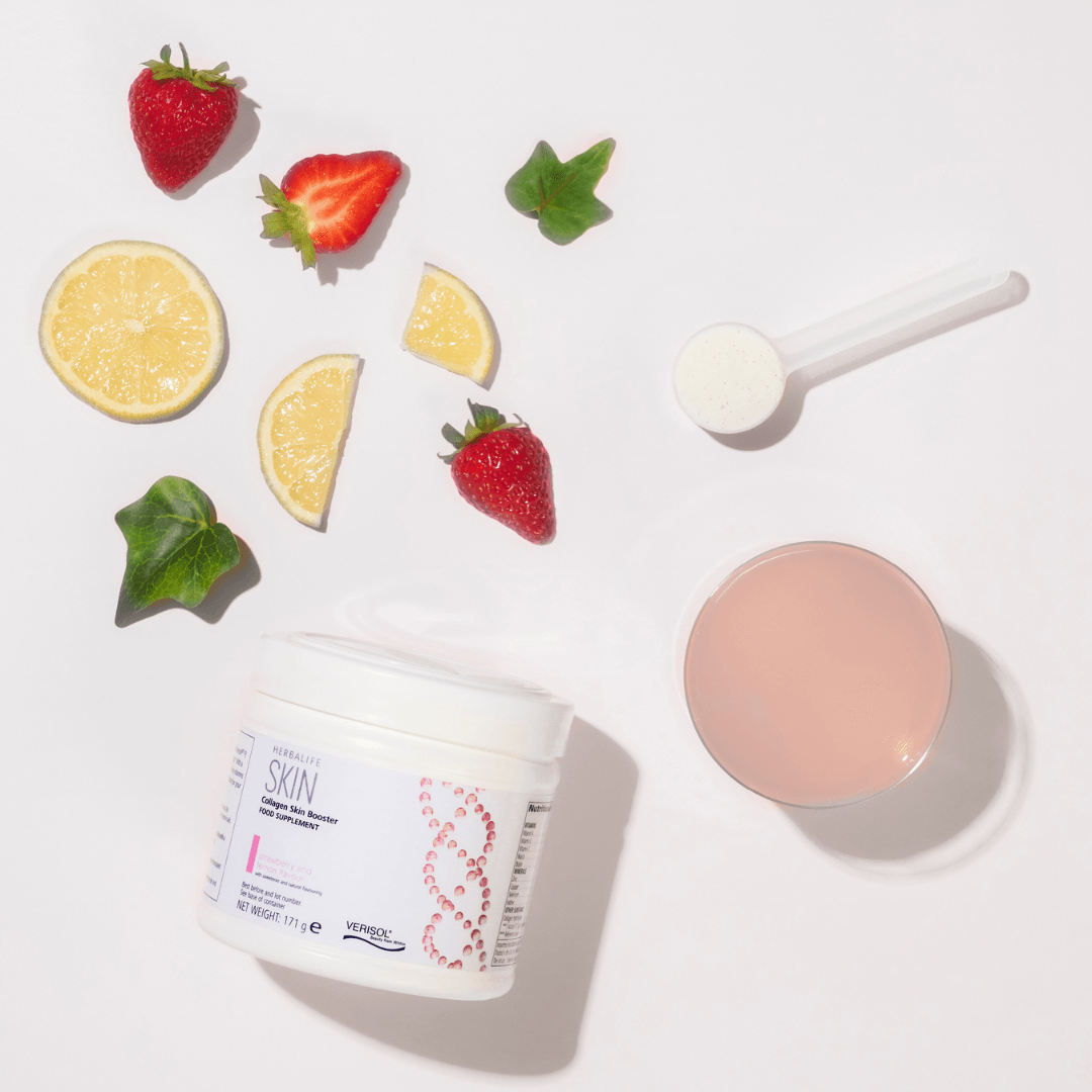 En boks Herbalife SKIN Collagen Booster ligger på et bord sammen med et glass drikkeklar kollagen, en måleskje og litt frukter. 