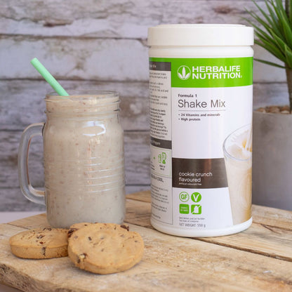 Herbalife Formula 1 shakemix med Cookie Crunch-smak og en Herbalife-shake laget som smoothie servert i et glassgrus. Foran ligger tre sjokoladecookies.