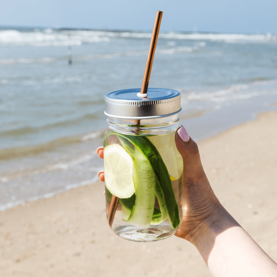 En hånd holder opp et krus med Herbalife aloeverajuice på ei strand. I glasset er det skivet sitron og agurk.