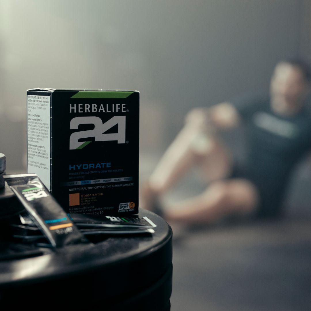 En pakke Herbalife H24 Hydrate isotonisk sportsdrikk og to løse porsjonsposer står på en stabel vektskiver. Utydelig i bakgrunnen ses en mann i treningstøy. 