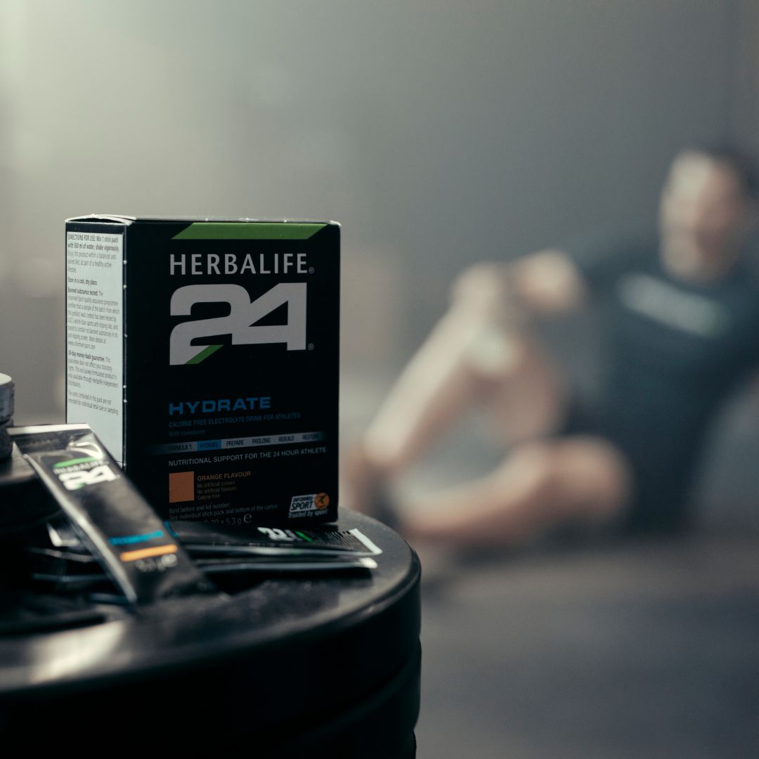 En pakke Herbalife H24 Hydrate isotonisk sportsdrikk og to løse porsjonsposer står på en stabel vektskiver. Utydelig i bakgrunnen skimtes en mann i treningstøy.  