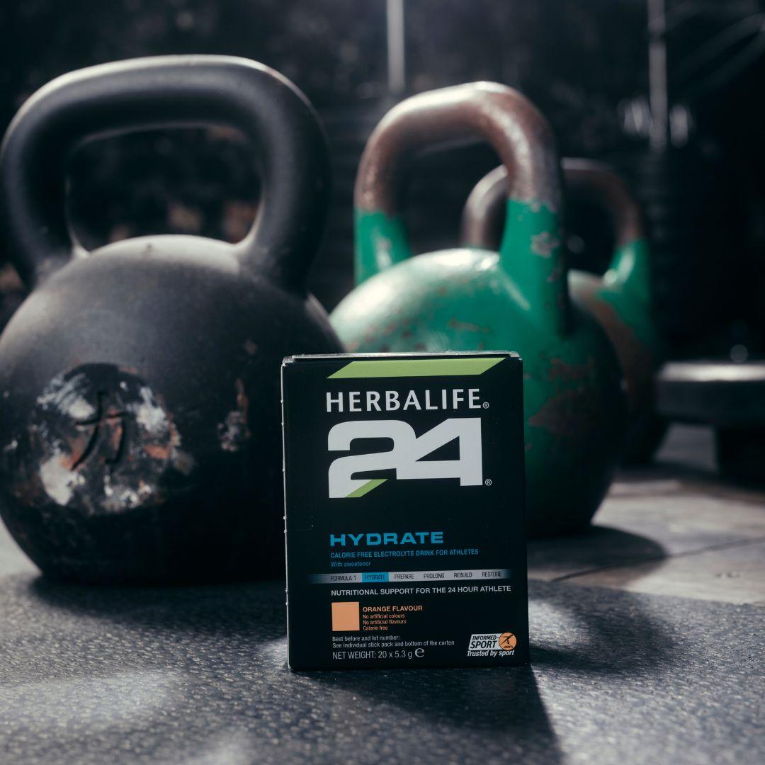 En pakke Herbalife H24 Hydrate isotonisk sportsdrikk står på gulvet foran noen kettlebells. 