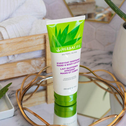 Tube med Herbalife Herbal Aloe Everyday Soothing Hand & Body Lotion på glassfat med gullkant på marmorbord. Aloe vera-plante, oppskårne aloevera-blad og trekasse med håndklær i bakgrunnen. 