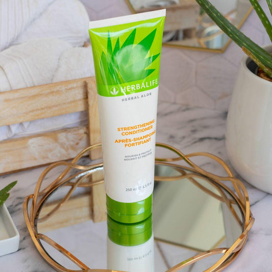 Herbalife Herbal Aloe Strengthening Conditioner-tube på glassfat med gullkant på marmorbord. Aloe vera-plante og oppskårne aloevera-blad, trekasse med håndklær og heksagonalt speil i bakgrunnen. 