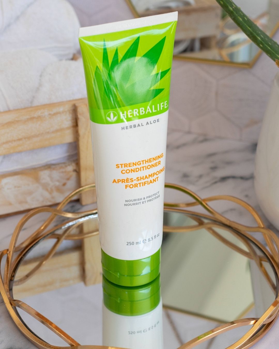Herbalife Herbal Aloe Strengthening Conditioner-tube på glassfat med gullkant på marmorbord. Aloe vera-plante og oppskårne aloevera-blad, trekasse med håndklær og heksagonalt speil i bakgrunnen. 