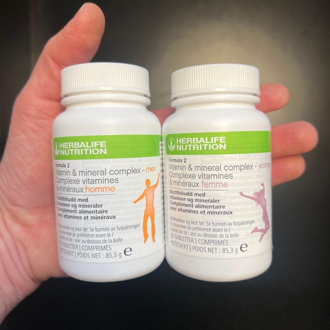 To bokser Herbalife Formula 2 Multivitamin, en versjon for kvinne og en for mann i hånda. Boksene har påskriften Kosttilskudd med vitaminer og mineraler. 