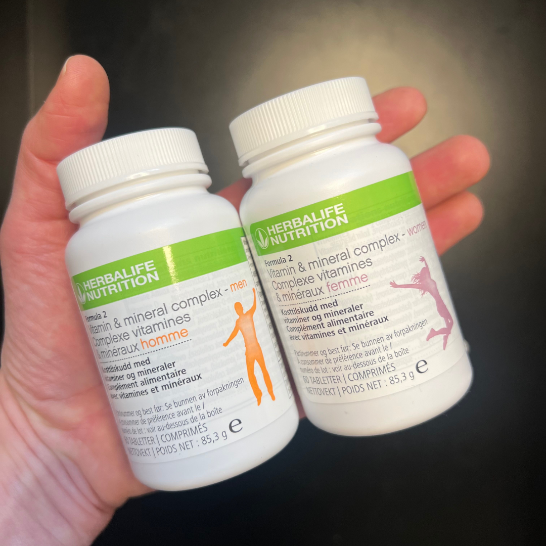 To bokser Herbalife Formula 2 Multivitamin, en versjon for kvinne og en for mann i hånda. Boksene har påskriften Kosttilskudd med vitaminer og mineraler. 