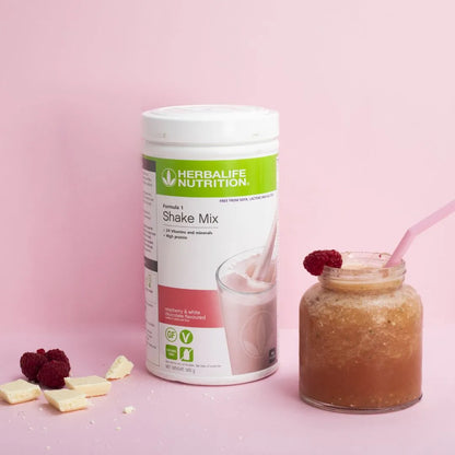 En boks Herbalife Formula 1 soyafri shakemix med Raspberry & White Chocolate-smak og en bringebærsmoothie servert i et glasskrus med et rosa sugerør, pyntet med bringebær. Noen biter av hvit sjokolade og hele bringebær ligger på bordet.