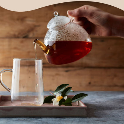 Rykende varm Herbalife Instant Urtete helles fra en glasskanne i en gjennomsiktig kopp, på et fat med dekorativer blader og blomst ved siden av. Perfekt for en sunn og oppkvikkende start på dagen. 