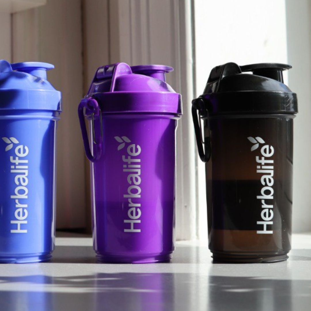 Tre Herbalife Smartshake-flasker i blå, lilla og svart, hver med hvit Herbalife-logo og karabinkrok, plassert på et bord ved et vindu.