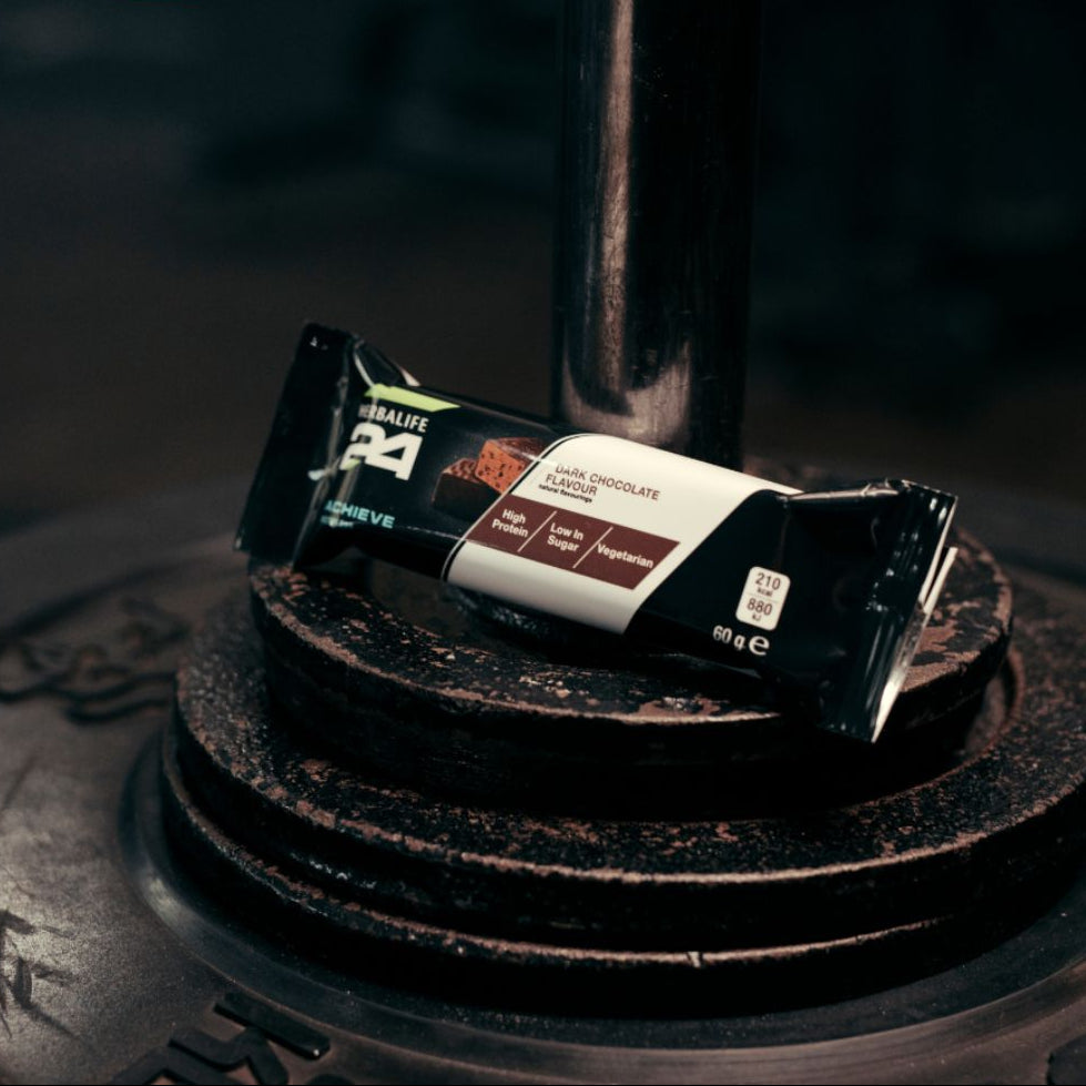 Herbalife H24 Achieve Protein Bar i smaken mørk sjokolade, plassert på vektskiver - perfekt snack etter trening med høyt proteininnhold og lavt sukkerinnhold.