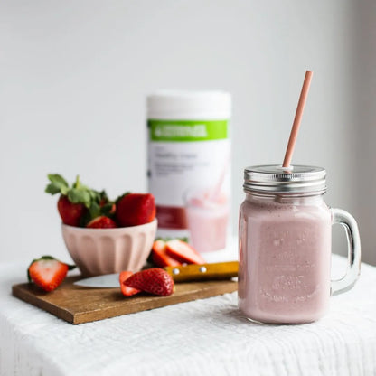 En Herbalife jordbær-shake i et glasskrus, noen jordbær i en skål og i bakgrunnen skimtes en boks Herbalife Formula 1 shakemix strawberry. 