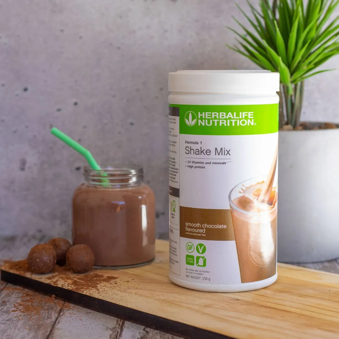 En boks Herbalife Formula 1 shakemix med Smooth Chocolate-smak og en sjokoladeshake i et glasskrus sammen med tre sjokoladekuler overstrødd med kakaopulver.