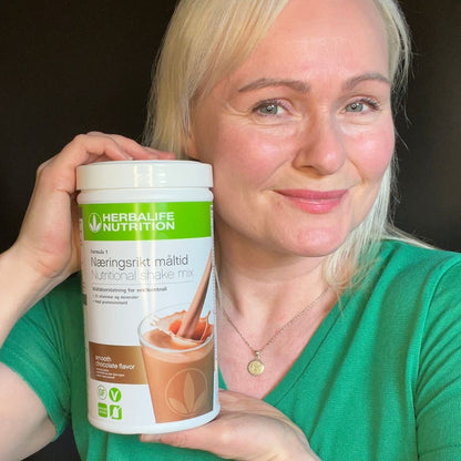 En boks Herbalife Formula 1 shakemix med Smooth Chocolate-smak og Helga som viser den frem. 