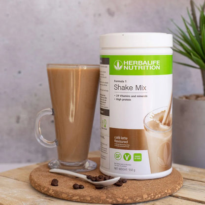 Boks Herbalife Formula 1 shakemix med Caffe Latte-smak på ei trefjøl. Ved siden av står et glasskrus med kaffelatte-shake, og foran ligger en hvit teskje med kaffebønner.